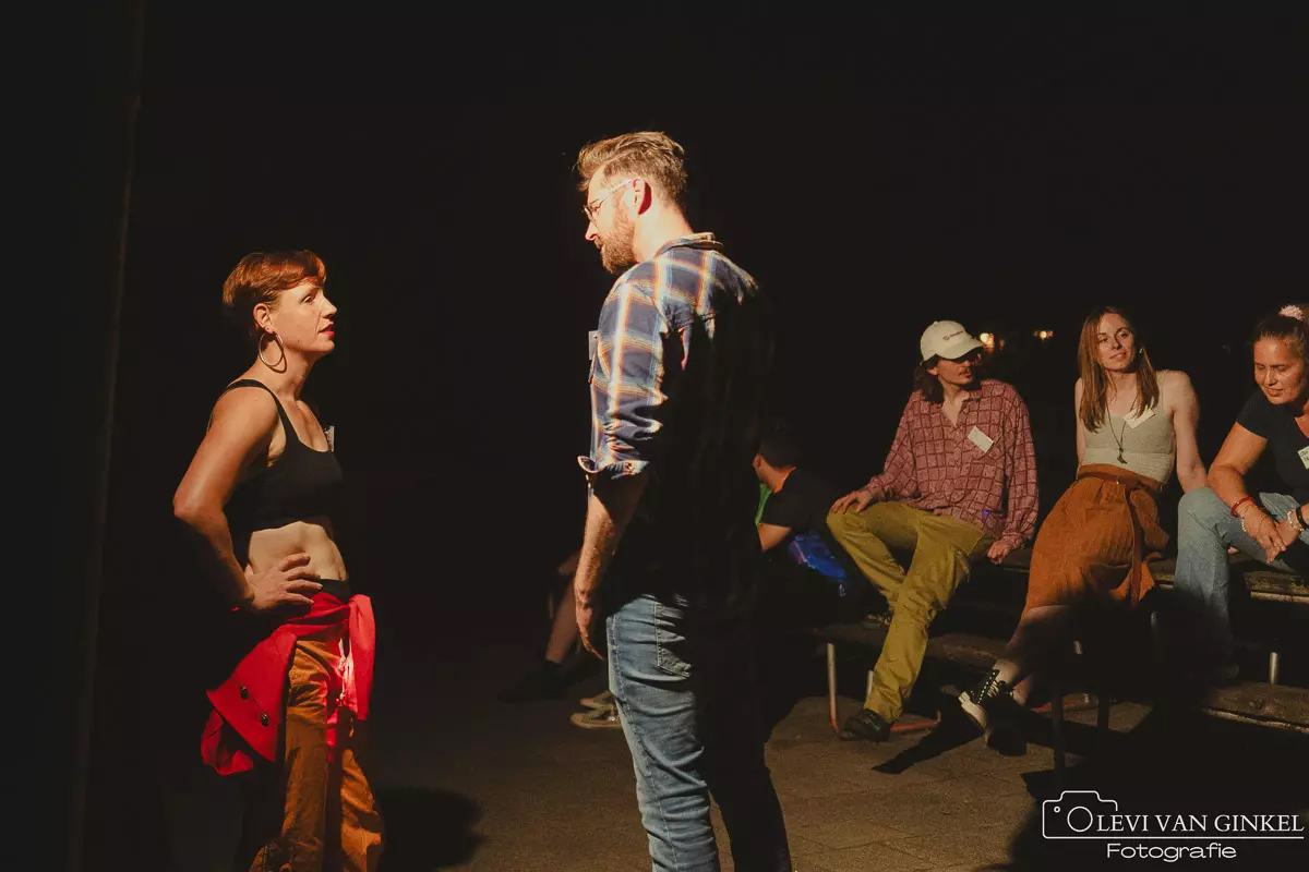 Twee deelnemers van een short larp praten met elkaar terwijl diverse anderen toekijken vanuit het donker.