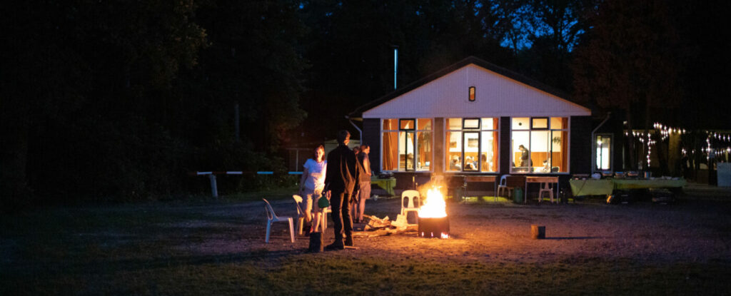 Deelnemers staan rond een kampvuur te praten met het locatiegebouw op de achtergrond.